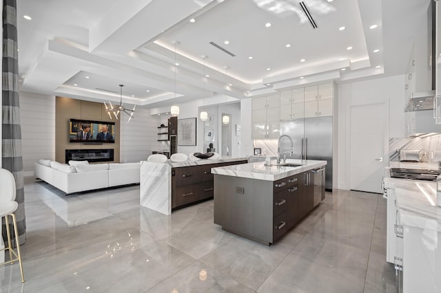 kitchen with a kitchen island with sink, a raised ceiling, dark brown cabinetry, a large fireplace, and white cabinetry