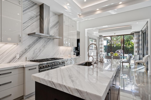 kitchen with wall chimney range hood, white cabinets, light stone counters, and an island with sink