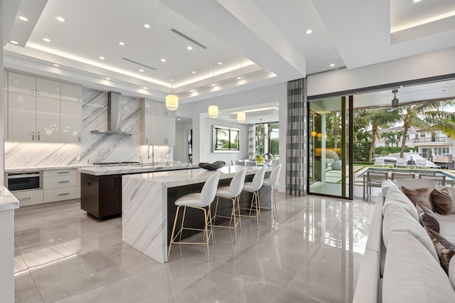 kitchen featuring wall chimney exhaust hood, a tray ceiling, pendant lighting, a center island, and white cabinetry