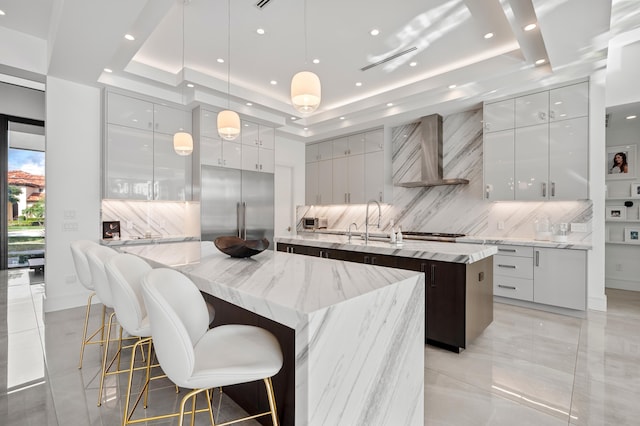 kitchen featuring light stone countertops, appliances with stainless steel finishes, a raised ceiling, wall chimney range hood, and a large island