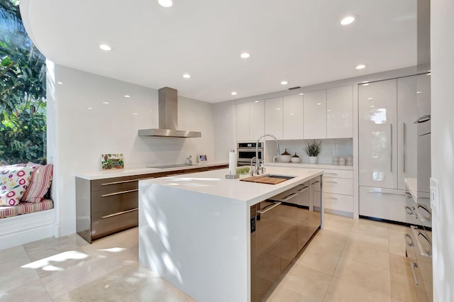 kitchen with wall chimney exhaust hood, stainless steel appliances, sink, white cabinets, and an island with sink