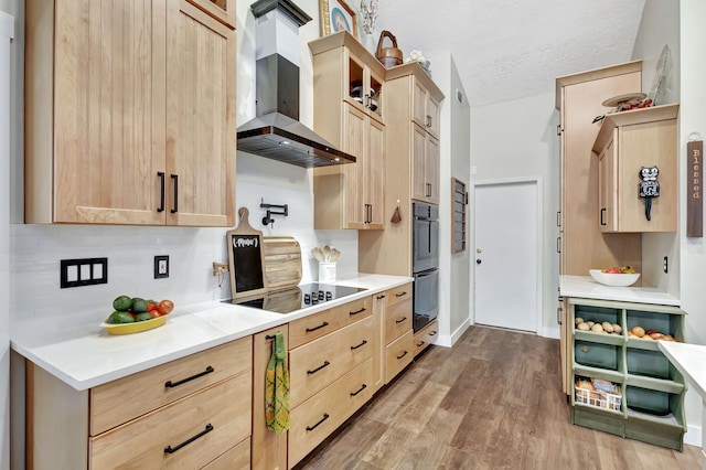 kitchen featuring multiple ovens, a textured ceiling, wall chimney range hood, light brown cabinets, and hardwood / wood-style flooring