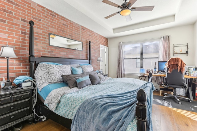 bedroom with a tray ceiling, ceiling fan, hardwood / wood-style floors, and brick wall