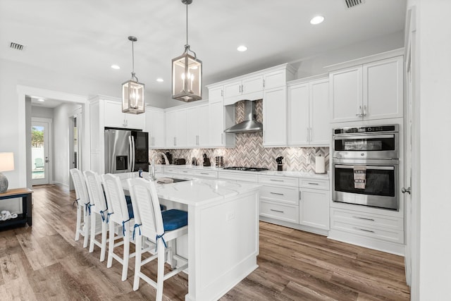 kitchen featuring stainless steel appliances, wall chimney range hood, pendant lighting, white cabinetry, and an island with sink