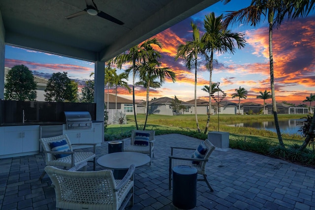 patio terrace at dusk featuring an outdoor kitchen, a water view, sink, ceiling fan, and a grill