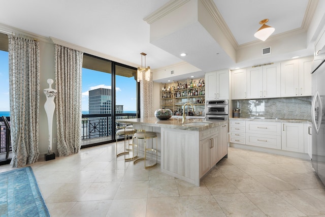 kitchen featuring ornamental molding, a kitchen island with sink, a wall of windows, white cabinetry, and hanging light fixtures