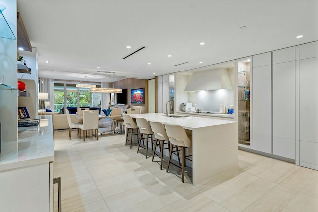 kitchen with a large island with sink, a kitchen bar, sink, white cabinetry, and wall chimney range hood