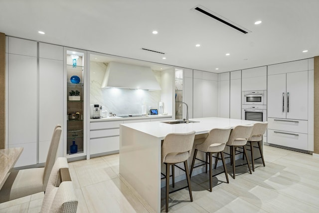 kitchen with an island with sink, a breakfast bar, wall chimney exhaust hood, white cabinets, and sink