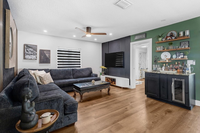 living room featuring beverage cooler, indoor bar, ceiling fan, a textured ceiling, and light hardwood / wood-style flooring