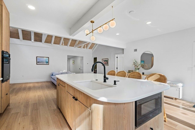 kitchen with a kitchen island with sink, black appliances, sink, light wood-type flooring, and decorative light fixtures