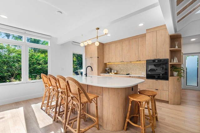 kitchen featuring a kitchen island with sink, hanging light fixtures, light brown cabinetry, tasteful backsplash, and black oven