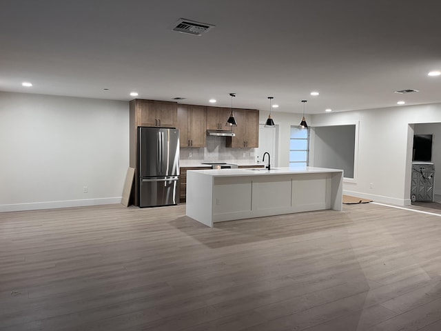 kitchen featuring hanging light fixtures, sink, light wood-type flooring, an island with sink, and stainless steel fridge