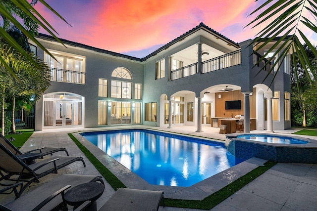 back house at dusk featuring a patio area, a swimming pool with hot tub, a balcony, and french doors