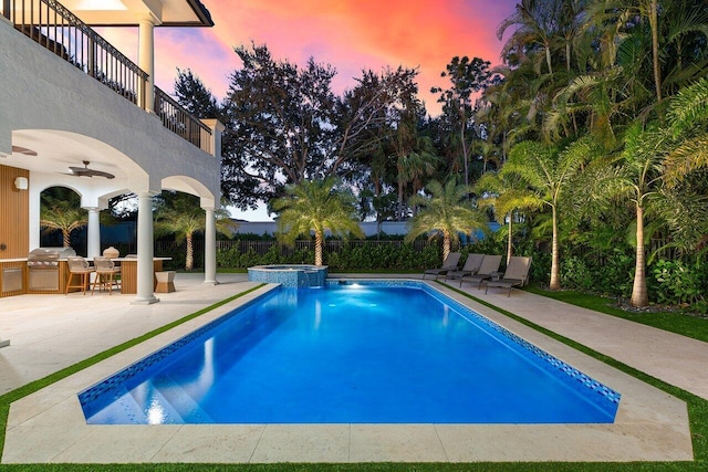 pool at dusk with an outdoor kitchen, ceiling fan, a patio, and an in ground hot tub