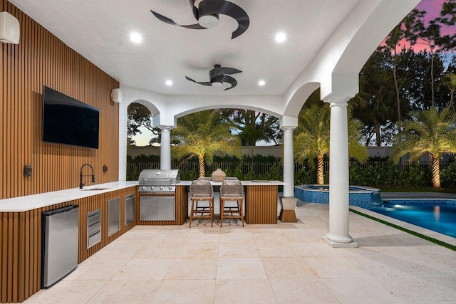 patio terrace at dusk featuring area for grilling, ceiling fan, a wet bar, and a pool with hot tub