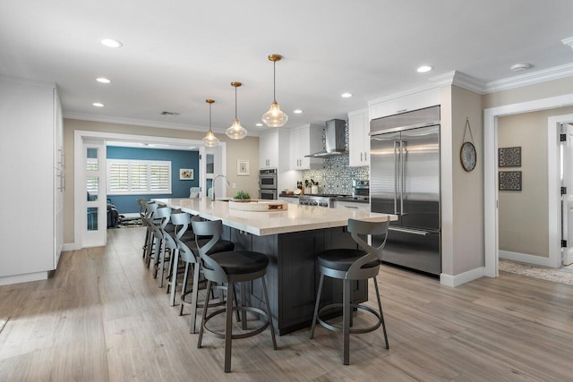 kitchen with wall chimney exhaust hood, pendant lighting, an island with sink, white cabinetry, and appliances with stainless steel finishes