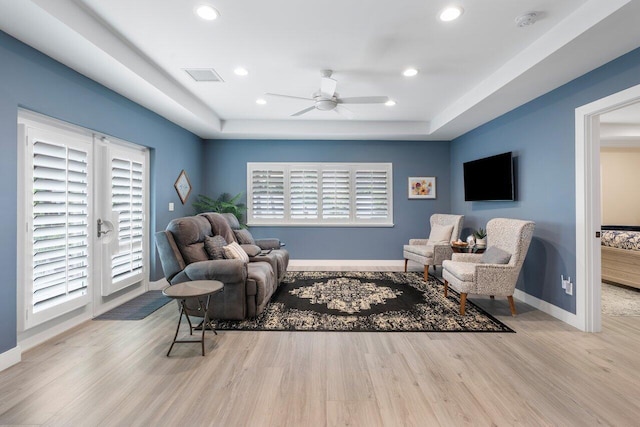 living area with ceiling fan, light hardwood / wood-style flooring, a raised ceiling, and plenty of natural light