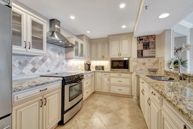 kitchen with wall chimney range hood, sink, appliances with stainless steel finishes, cream cabinetry, and light stone counters