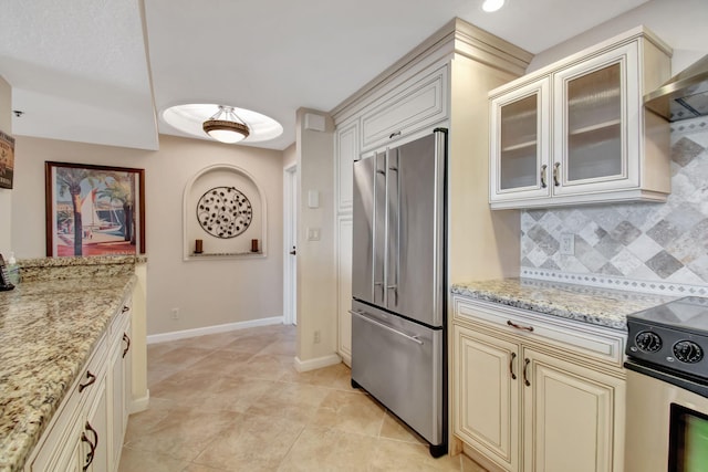 kitchen featuring cream cabinets, wall chimney exhaust hood, decorative backsplash, light stone counters, and stainless steel appliances