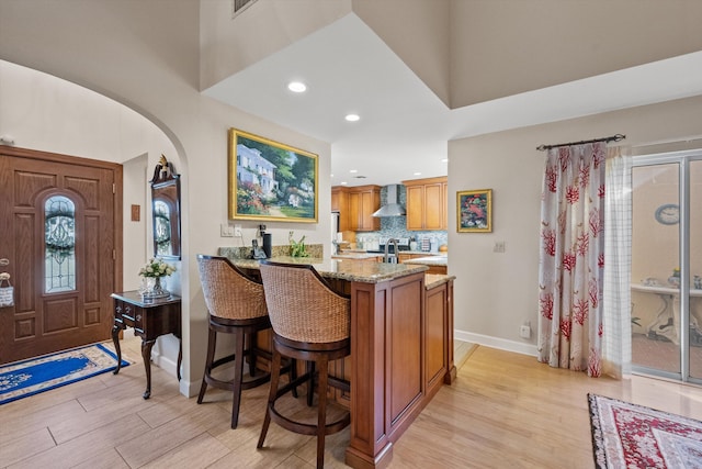 bar with light hardwood / wood-style floors, stainless steel stove, light stone counters, wall chimney range hood, and tasteful backsplash