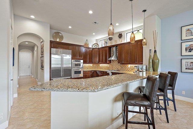 kitchen with light stone counters, a peninsula, stainless steel appliances, backsplash, and wall chimney exhaust hood