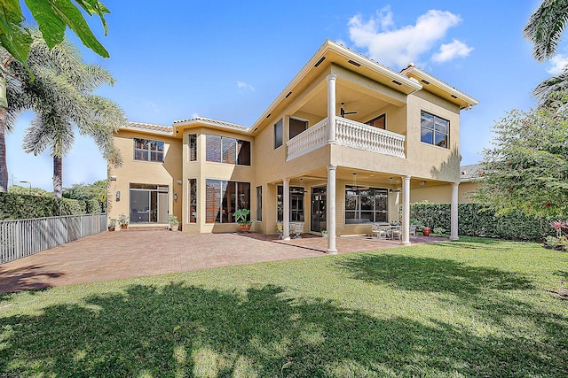 rear view of property with ceiling fan, a balcony, fence, a lawn, and stucco siding