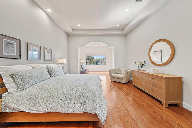 bedroom featuring arched walkways, light wood-style flooring, recessed lighting, baseboards, and a tray ceiling