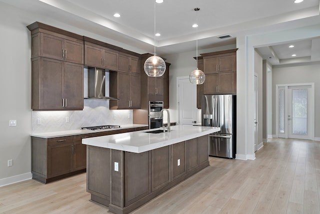 kitchen featuring appliances with stainless steel finishes, decorative light fixtures, a raised ceiling, wall chimney exhaust hood, and light wood-type flooring