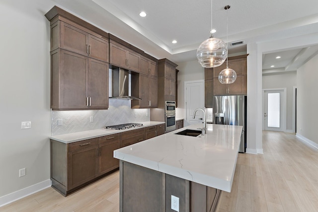 kitchen with sink, an island with sink, stainless steel appliances, light stone countertops, and wall chimney range hood