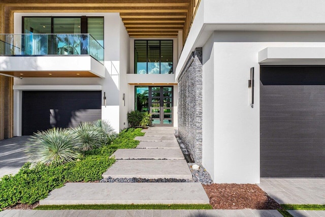 view of exterior entry with french doors, an attached garage, a balcony, and stucco siding