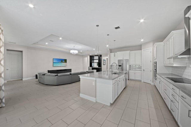 kitchen featuring tasteful backsplash, wall chimney exhaust hood, black electric cooktop, white cabinetry, and an island with sink