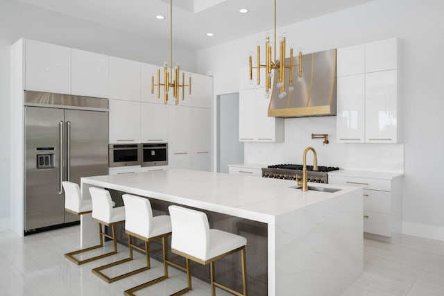 kitchen with appliances with stainless steel finishes, a kitchen island with sink, exhaust hood, decorative light fixtures, and white cabinets