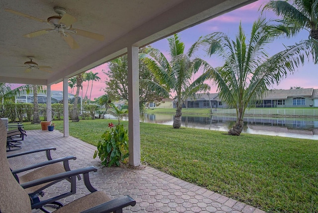 patio terrace at dusk featuring ceiling fan, a water view, and a yard