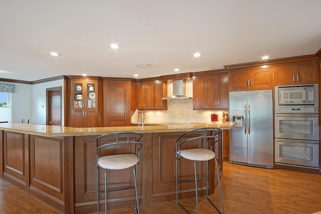 kitchen featuring appliances with stainless steel finishes, tasteful backsplash, wall chimney exhaust hood, light hardwood / wood-style floors, and an island with sink