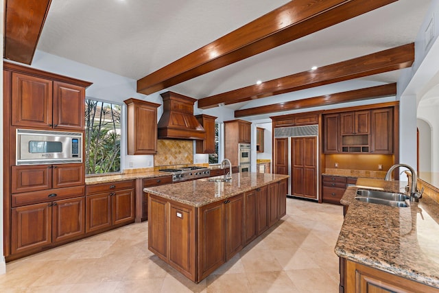 kitchen with light stone counters, custom exhaust hood, sink, beam ceiling, and built in appliances