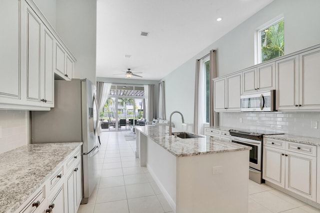 kitchen with light stone counters, a center island with sink, appliances with stainless steel finishes, ceiling fan, and decorative backsplash