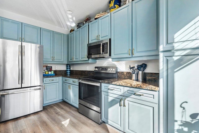 kitchen with blue cabinets, dark stone countertops, a textured ceiling, appliances with stainless steel finishes, and light wood-type flooring