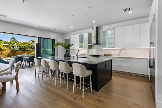 kitchen with wall chimney exhaust hood, a breakfast bar, a center island with sink, stone counters, and white cabinets