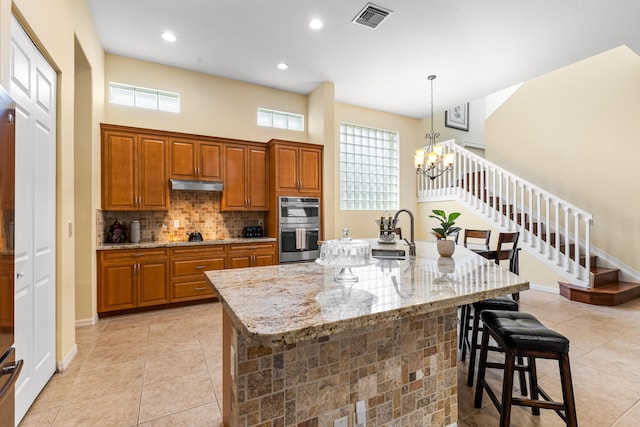 kitchen with a kitchen bar, stainless steel double oven, an island with sink, hanging light fixtures, and light stone counters