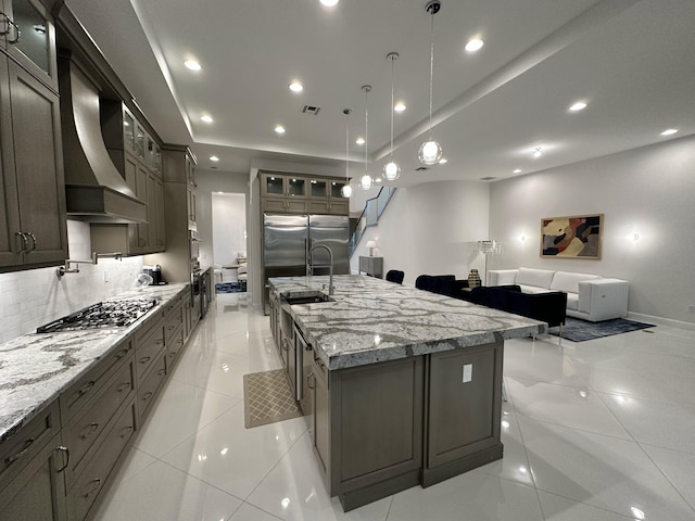 kitchen featuring a large island with sink, wall chimney exhaust hood, light tile patterned floors, decorative light fixtures, and stainless steel appliances