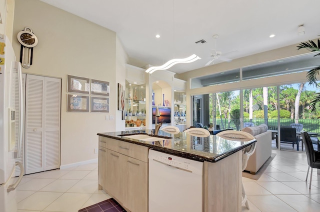 kitchen with dishwasher, light brown cabinets, a center island with sink, ceiling fan, and dark stone countertops