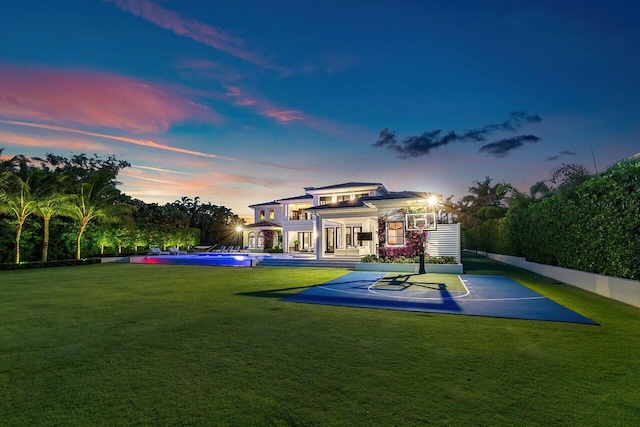 back house at dusk featuring a lawn and basketball hoop