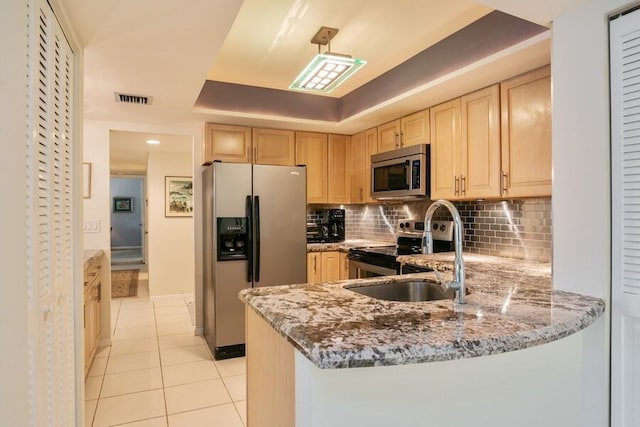 kitchen with kitchen peninsula, stainless steel appliances, a tray ceiling, light stone counters, and light brown cabinetry