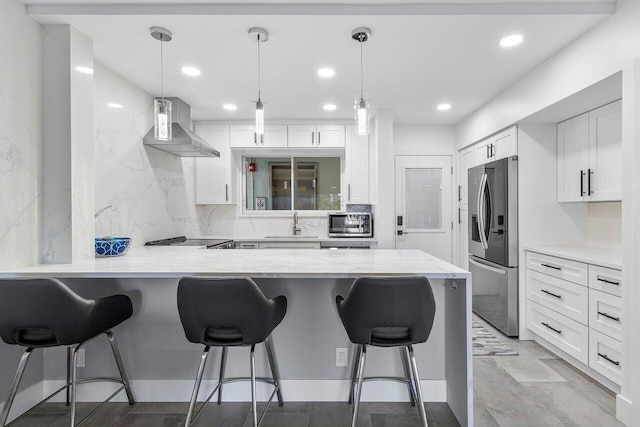 kitchen with wall chimney range hood, stainless steel refrigerator with ice dispenser, decorative light fixtures, white cabinetry, and a breakfast bar area