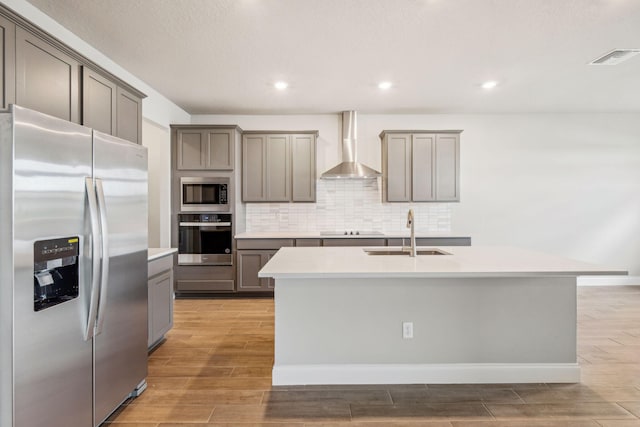 kitchen featuring wall chimney range hood, sink, a kitchen island with sink, backsplash, and stainless steel appliances