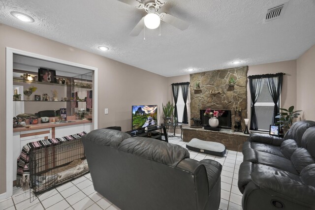 living room with a textured ceiling, a stone fireplace, ceiling fan, and light tile patterned flooring