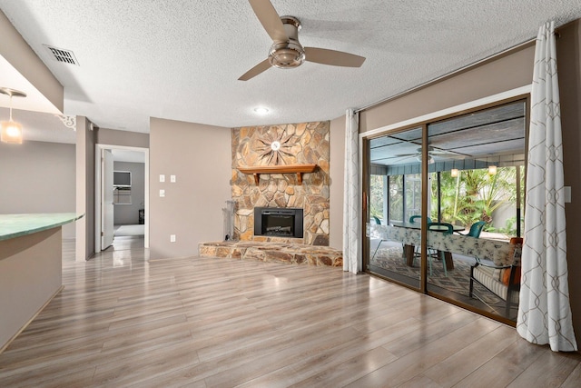 living room with ceiling fan, a stone fireplace, a textured ceiling, and light hardwood / wood-style flooring