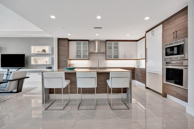 kitchen featuring built in appliances, a kitchen bar, white cabinetry, and wall chimney exhaust hood