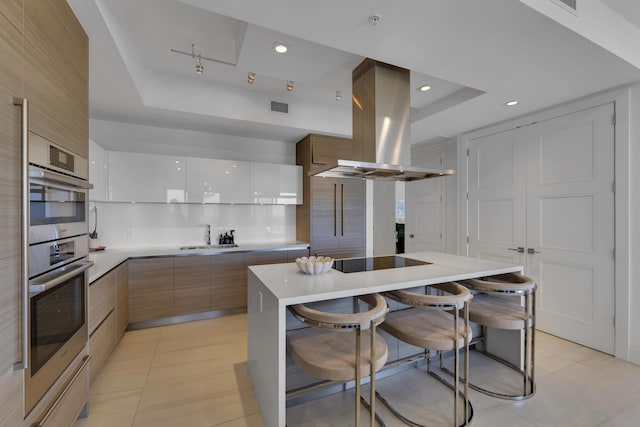 kitchen featuring a center island, island range hood, a tray ceiling, white cabinets, and a breakfast bar area