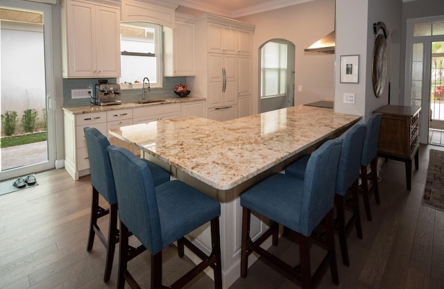 kitchen with white cabinets, wall chimney exhaust hood, sink, light stone counters, and a breakfast bar